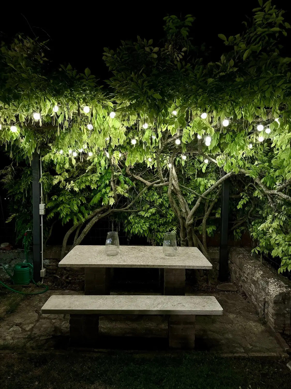 Stone table and benches under the wisteria pergola at night, lit by festoon lights